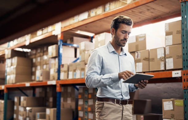 A man uses a tablet while standing in a warehouse filled with shelves of boxes.